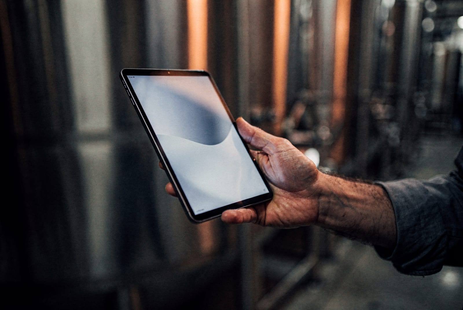 A brewer holding a slim tablet beside fermentation tanks.