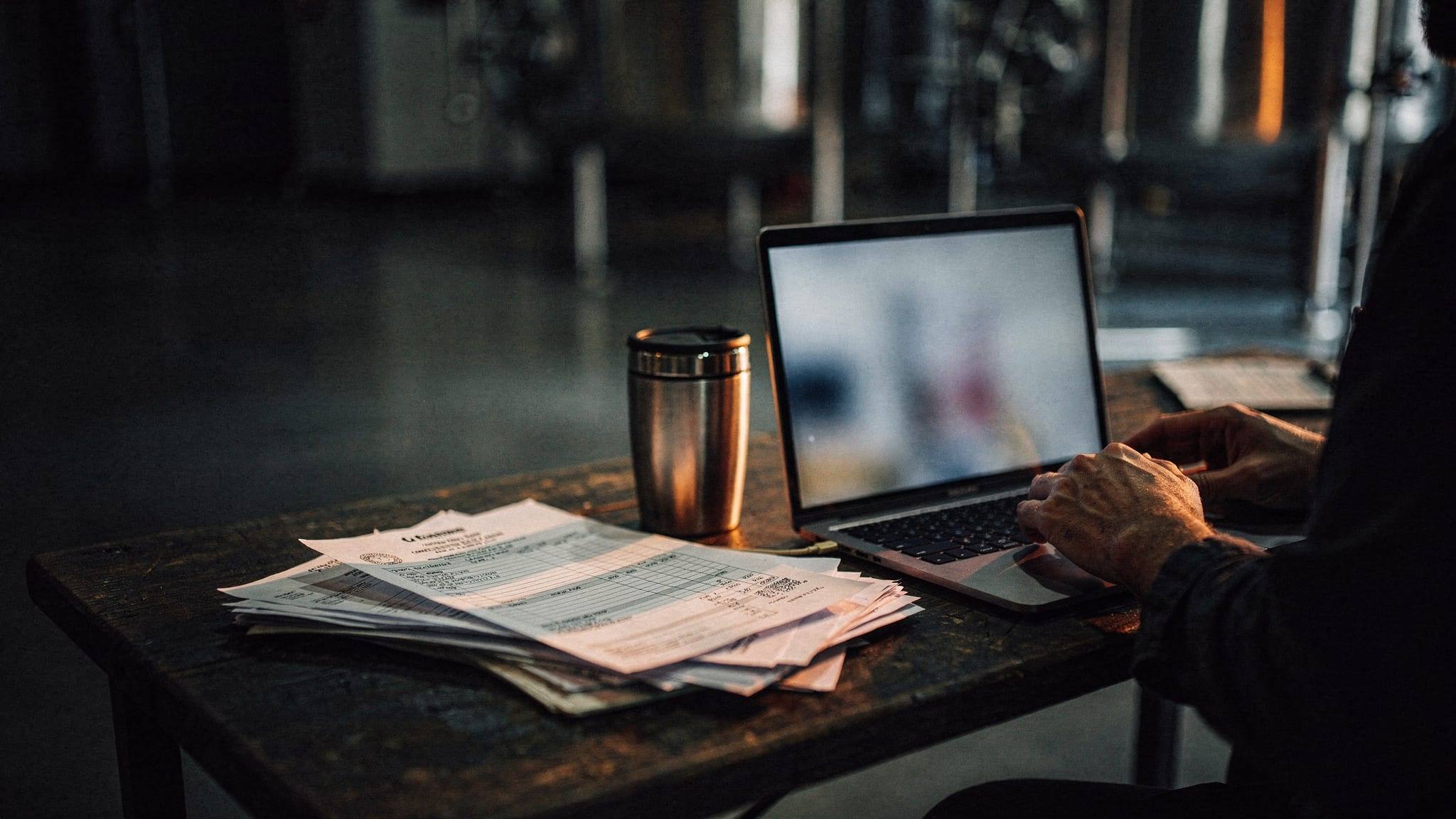 A laptop and a pile of paper purchase orders on a production office desk.