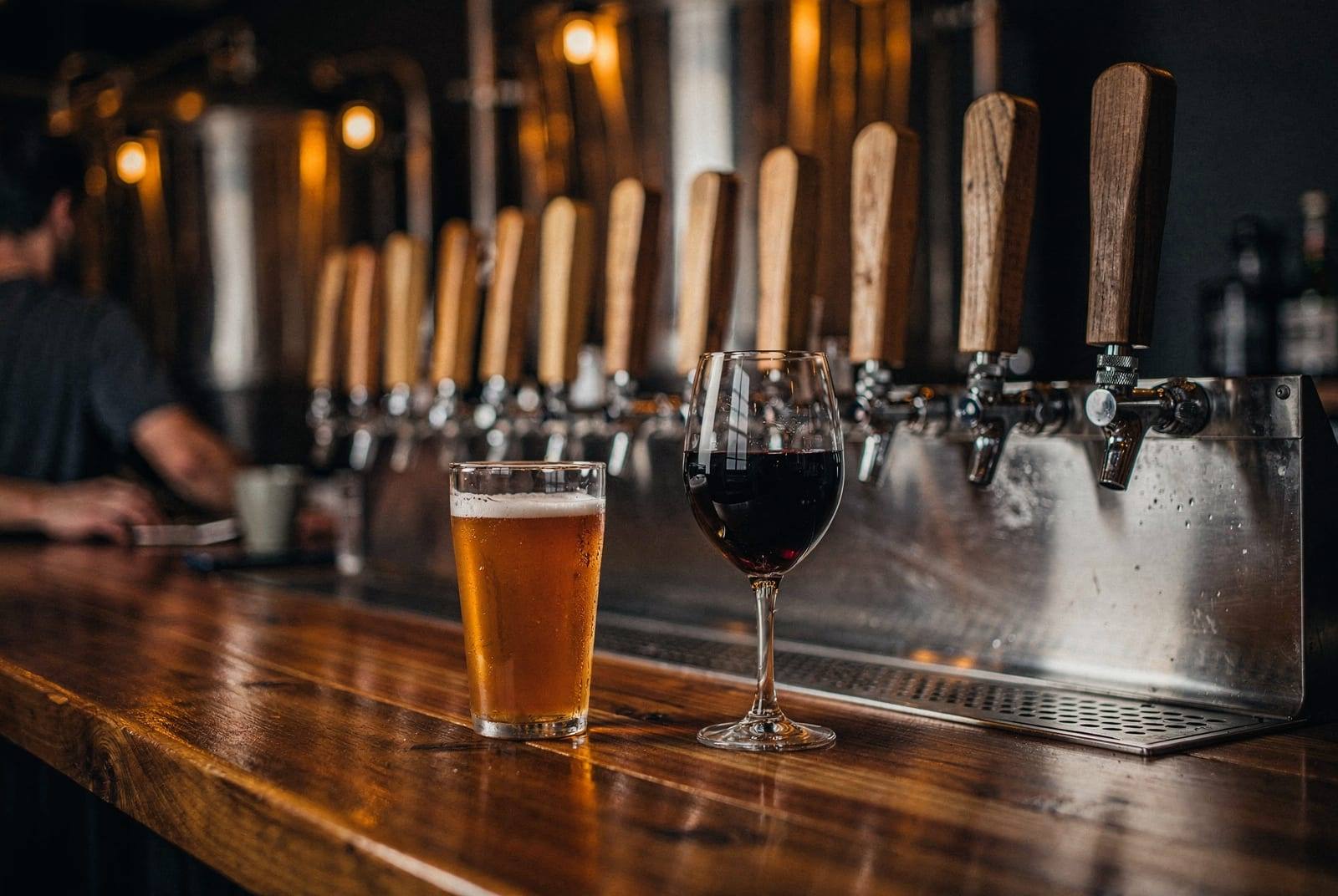 A glass of beer and a glass of wine on a warmly lit taproom bar.